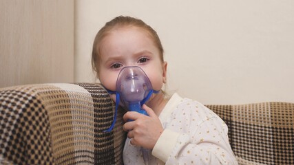 A cute little baby in a mask breathes through an inhaler. Home inhalation procedure. The child is receiving respiratory therapy with a nebulizer. Treat kid from coronavirus at home. A little girl