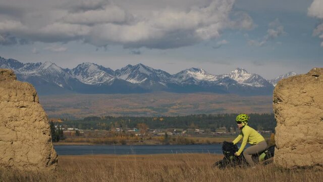 The woman travel on mixed terrain cycle touring with bikepacking. The traveler journey with bicycle bags. Sport bikepacking, bike, sportswear in green black colors. Mountain snow capped, stone arch.