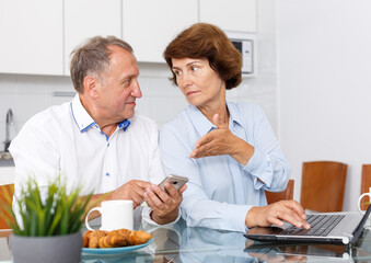 Mature man and woman using smartphone and working at laptop at home