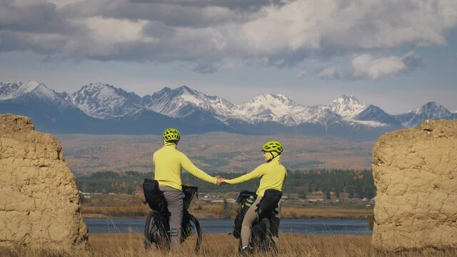 The man and woman travel on mixed terrain cycle bike touring with bikepacking. The two people journey with bicycle bags. Sport sportswear in green black colors. Mountain snow capped, stone arch.