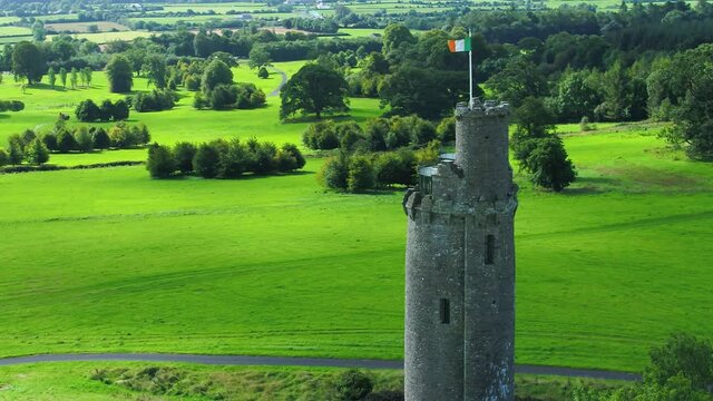 Irish flag blowing in wind a top tall castle tower in vast green landscape. Aerial