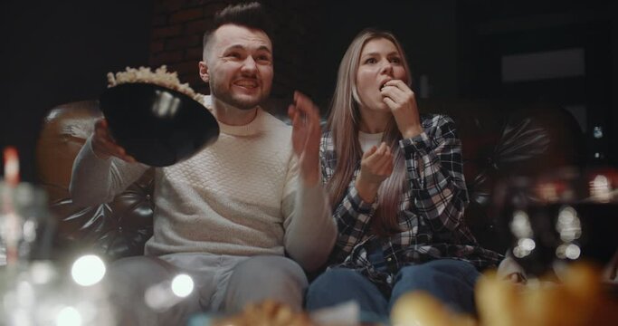 Excited millennials sitting on couch having fun watching football or basketball game on TV at home. Handsome bearded man cheering for favorite team screams and celebrates goal scatter popcorn