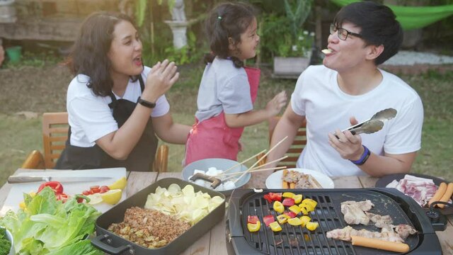 Happy Asian family barbecue grill in garden. Parents with little daughter enjoy cooking grilled meat and vegetable together at home. Father and mother with child girl kid having fun weekend activity.