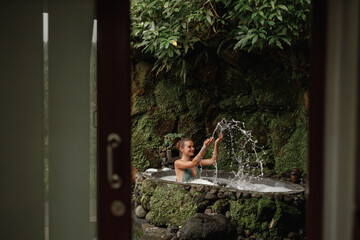 Young woman sit on outdoor bath tube, make splashes and enjoy time in spa. Organic skin care