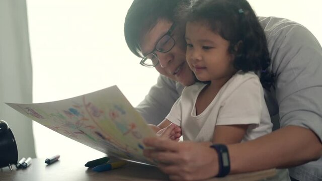Happy Asian family at home. Father teach little daughter using color pencil drawing and painting on paper book in living room. Parent with cute child girl kid having fun leisure homeschooling together