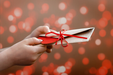 A human hand holds and gives a craft envelope and a figurine of a red heart. Holiday gift. Romantic love letter for Valentine's day concept. Bokeh background. Place for text