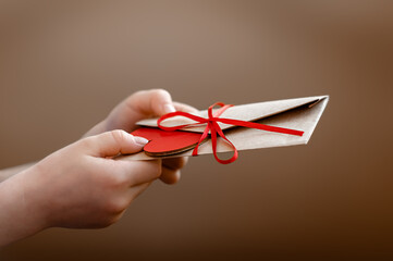 A human hand holds and gives a craft envelope and a figurine of a red heart. Holiday gift. Romantic...