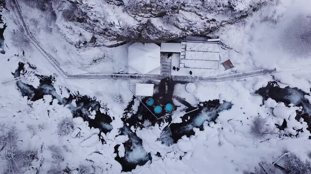 Hot springs in the snowy forest and mountains. Top view from the drone on the white gorge with the river. There are barrels of radon water. There is steam, people are bathing. Almarasan, Almaty.
