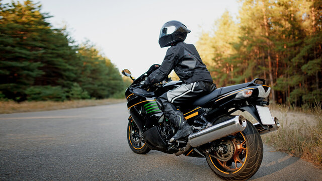 Motorcyclist alone on the road in a helmet and leather protective equipment looks to the side, sitting on a fast sports motorcycle on a forest background