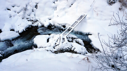 A clear mountain river runs through a snowy gorge. There is a metal bridge covered with snow. Tall spruce trees grow on the slopes of the mountains. There is steam from the river. Almarasan, Almaty