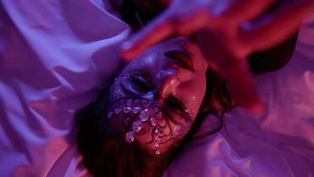 Face a woman with beautiful makeup, a pattern and with rhinestones on her face poses in front of the camera in the studio lying on the bed. Makes a motion with his hands.