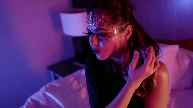 A female model with a beautiful makeup, a pattern and with rhinestones on her face poses beautifully in front of the camera in the studio sitting on the bed.
