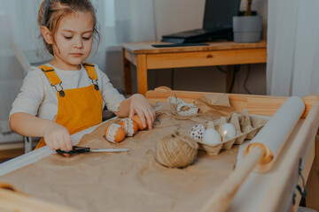 Fototapeta na wymiar Baby girl decorating easter eggs on white table. Kid decorating eggs with kraft paper, lace and rope. Child cuts the rope with black scissors.