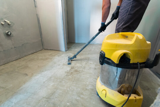 The builder cleans the floor from dust with a construction vacuum cleaner.