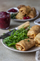 Fototapeta na wymiar Homemade stuffed thin pancakes or crepes with meat, spicy cranberry sauce and arugula, light stone background. Traditional Russian cuisine for Maslenitsa. Shrove Tuesday.