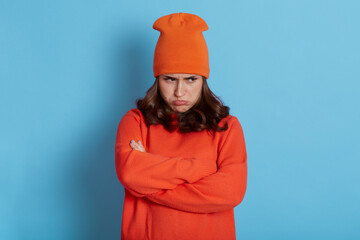 Dark haired caucasian woman stands with arms folded over body, looks from under forehead with pout...