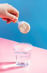 Female hand pouring protein powder into glass of water on blue pink background.