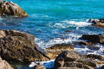 Turquoise sea stone beach, breaking waves in bright sunlight. Colorful bright sea background. The concept of summer, vacation, travel. The purest clear sea water, large stones on the beach close-up