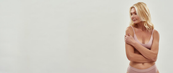 Body acceptance. Beautiful middle aged blonde woman in lingerie hugging herself, looking away while posing isolated over grey background, copy space for text