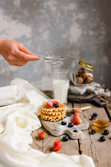 A woman's hand sprinkles icing sugar on homemade waffles. The icing sugar goes to the fresh perfect waffles. Idea for breakfast or dessert.