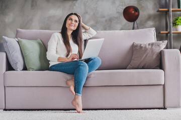 Photo of positive minded girl sit sofa barefoot hold laptop wear eyewear white pullover in living room home indoors