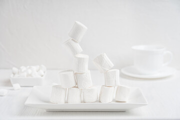 Pile of white sweet marshmallows levitating or balancing on top of one another on rectangular plate on white wooden background. Horizontal orientation image. Flying food concept