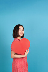 joyful, cheerful young asian woman in red dress holding big red paper heart like fan isolated on blue background.