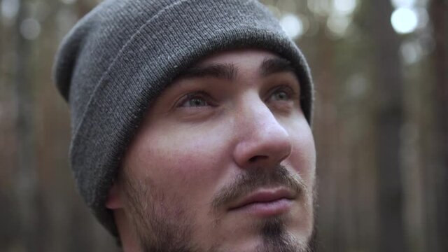 Close-up Portrait of a Young Man in a Knitted Hat on a Blurred Forest Background. Travel Concept, Hicking. Satisfied Traveler Guy Stands in Picturesque Place of Wildlife and Looks at Autumn Landscape.