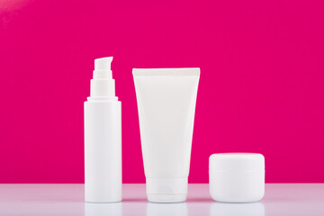 Fototapeta na wymiar Minimalistic still life with three white glossy cream jars on white table against pink background. Concept of daily skin or hair care.