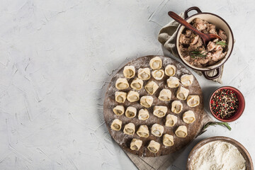 Lots of ready-to-cook pork dumplings on a large wooden board with flour and meat in bowls.