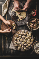 Lots of ready-to-cook pork dumplings on a large wooden board with flour and meat in bowls on a dark wooden background.