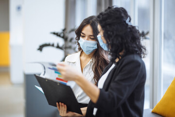 Two women checking charts and discussing business trends in the office. Females colleagues wearing medical masks sit on the couch at a working place. Coronavirus prevention. Colaboration concept.