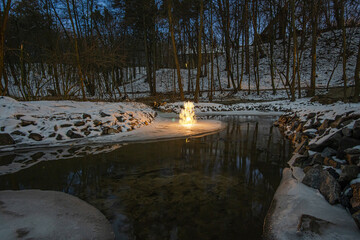 Bonfire on the shore of a forest stream or river  in winter at night with silhouettes of low-rise houses in the background.