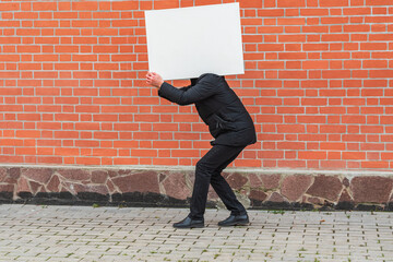 A man carries on his shoulder a blank white canvas for painting against a red brick wall 