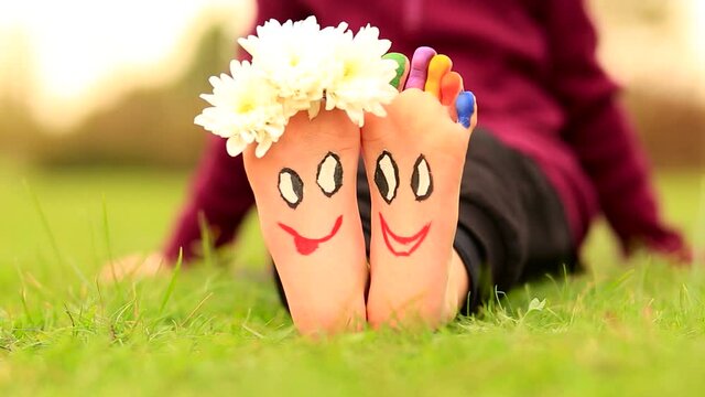 Little child girl sits on green grass with painted feet with funny comic faces 