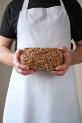 Close-up at bakers hands holding a multi-seeded loaf of sourdough whole grain bread in front of him.