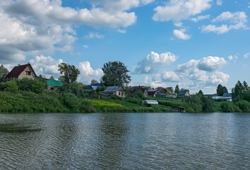 Fototapeta na wymiar Village by the lake on a summer sunny day. By the lake, there are picturesque wooden houses with vegetable gardens and greenhouses, a blue sky with white cumulus clouds 