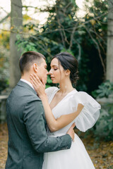 Naklejka premium attractive bride in a delicate fashionable wedding dress kisses the groom in a gray suit near stone columns in a tropical garden on a warm summer day.