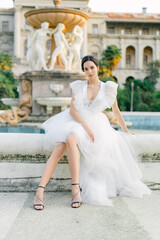 Portrait of Beautiful bride in white fashion wedding dress. Outdoor romantic portrait of attractive brunette woman with hairstyle posing by fountain at park.