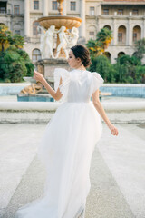 Gorgeous bride posing at fountain in park, wearing adorable bridal lace dress. Smiling young woman in wedding dress posing on camera