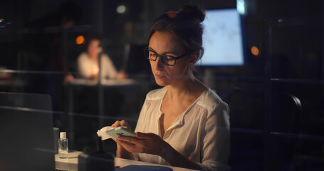 Young businesswoman disinfecting smartphone with sanitizer gel working late in office