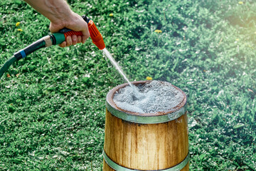Man cleans or fills vintage oak wood tub with clear water from hose on lush green lawn grass on sunny summer day close view