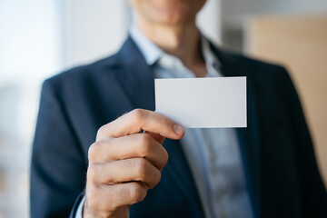 Man holding blank business card with copy space, standing in modern office. Mock up, advertisement concept 
