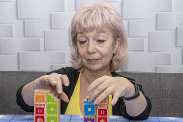 Portrait of an elderly woman 60-65 years old, who collects a wooden puzzle from cubes at the table.