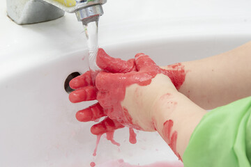 Fototapeta premium The girl washes her hands of red paint in the washbasin. Concept: children's educational games with paints, personal hygiene.