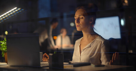 Businesswoman relaxing and meditating sitting at workplace in modern office at night