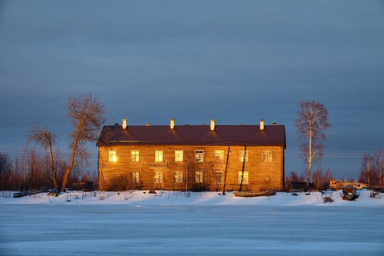 Two-storey log houses (barracks) on Northern river, village. North-Western Russia. Wooden architecture of early 20th century. Taking photo from frozen river, evening, low winter sun, long shadows