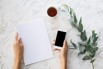 Female hands with blank magazine, mobile phone and cup of tea on light background