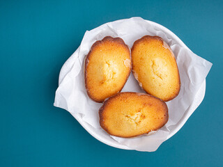 Lemon glaze madeleines on a plate placed on a blue light background. Madeleine - homemade traditional French small cookie. Seashell sweet cakes
