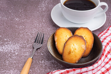 Madeleine homemade traditional French small cookies on black a dish and a coffee cup placed on a wooden background. Lemon glaze madeleines. Seashell sweet cakes